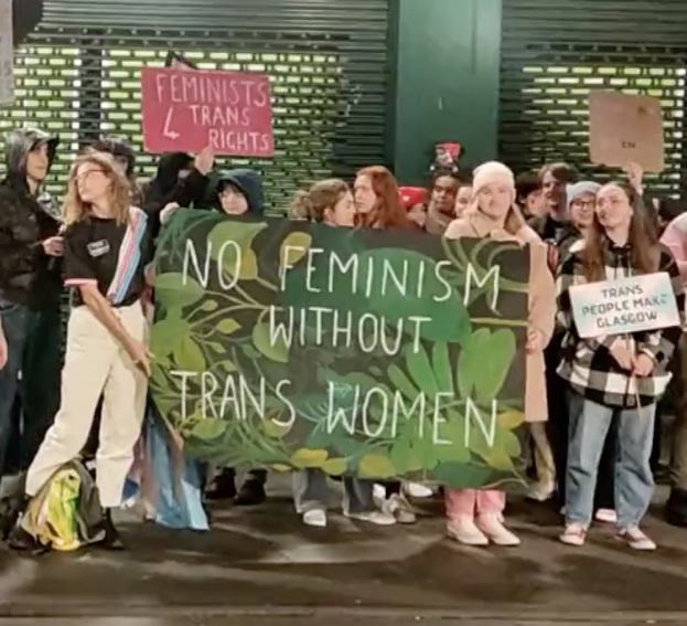 Gruppe von Menschen mit einem Banner, das "Kein Feminismus ohne Trans-Frauen" und Schilder in einer öffentlichen Fläche mit einer Wand und einem Rollladen im Hintergrund zeigt.