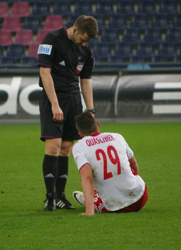 Ein Fußballer sitzt neben einem Schiedsrichter auf dem Boden in einem Stadion, beide tragen Sportkleidung, mit Schildern und Stühlen im Hintergrund.