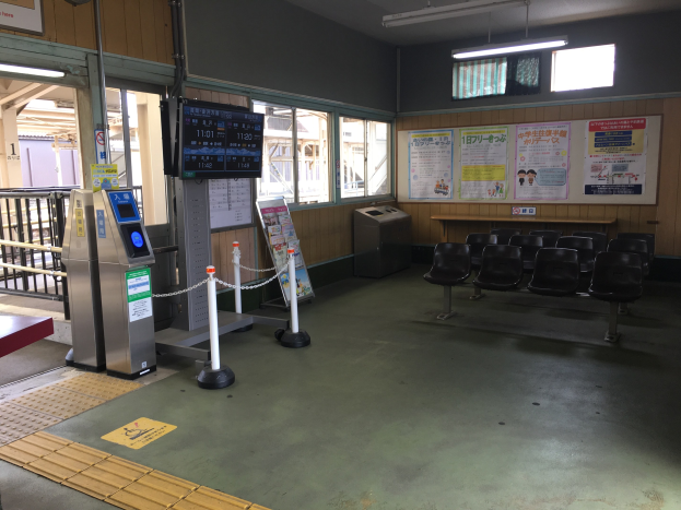 Ein belebter Bahnhofseingang mit Sitzgelegenheiten, einer Informations counter, Ticketautomaten, Kettenpollern, Informationsanzeigen, Wandplakaten, Deckenleuchten und Geländern.