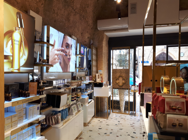 Innenansicht eines Kosmetikladens mit verschiedenen Produkten auf Gestellen, Bildschirmen an den Wänden, Deckenleuchten, einer Klimaanlage und Glastüren, die eine Person, ein Auto und ein Gebäude draußen zeigen.