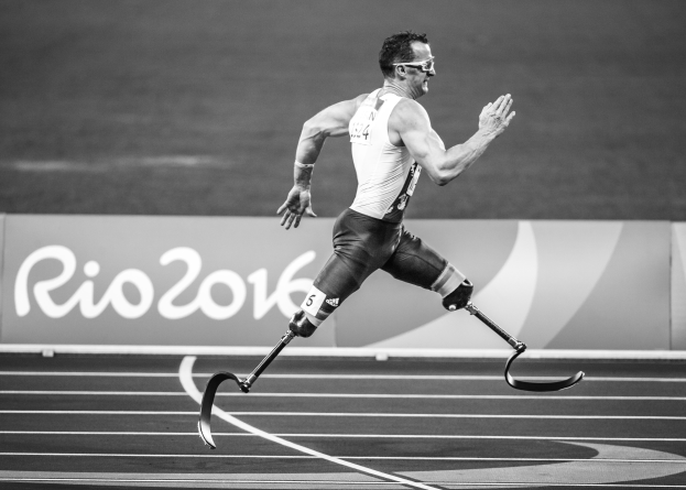 Ein Mann mit einer Prothese läuft auf einer Laufbahn, im Hintergrund ist ein Schild zu sehen mit der Aufschrift "Rio 2016 Paralympic Games", dargestellt in Schwarz-Weiß.