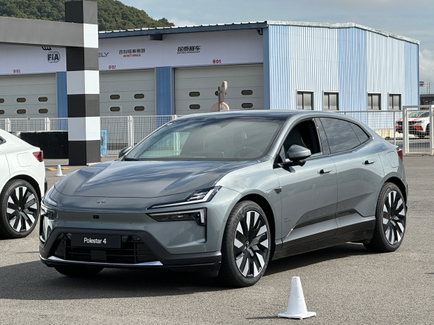 Zwei Elektroautos, ein Polestar 4 und ein Tesla Model X, nebeneinander vor einem Gebäude mit einem Verkehrskegel im Vordergrund geparkt. Im Hintergrund sind Geländer, Schuppen, Bäume und ein klarer blauer Himmel zu sehen.