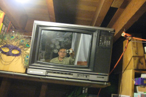 Ein Fernsehbildschirm zeigt eine Person umgeben von Pfauenfedern, Büchern und Schachteln.