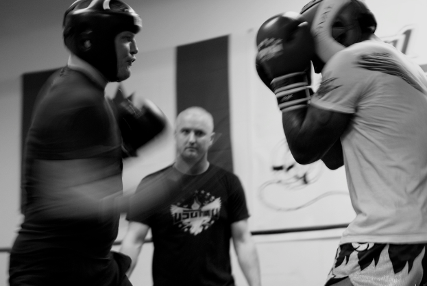 Zwei Boxer mit Helmen und Handschuhen kämpfen in einem Ring, wobei ein Schiedsrichter zwischen ihnen steht und ein Banner an der Wand im Hintergrund zu sehen ist.