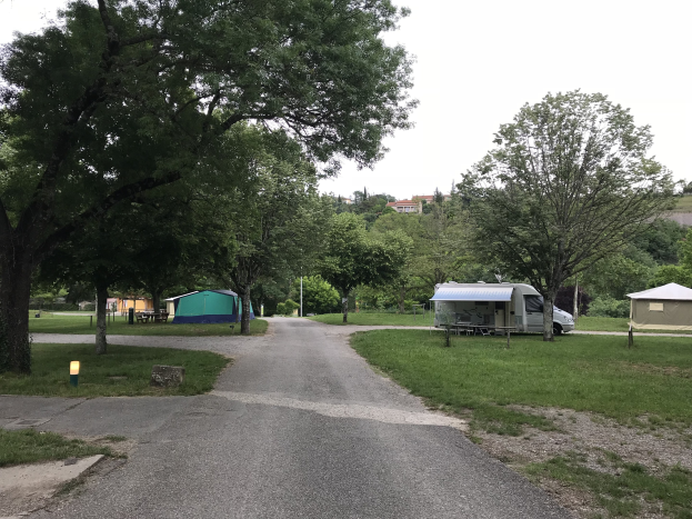 Campingplatz mit einem Auto neben einem Baum geparkt, grasigem Boden, verstreuten Zelten und Bänken, Bäumen und Gebäuden im Hintergrund unter einem klaren blauen Himmel.