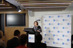 Eine Frau steht auf einer Bühne und spricht in ein Mikrofon, vor ihr sitzen Zuschauer, an der Wand hängt ein Banner, an der Holzwand steht ein Fernseher und an der Decke sind Lampen.