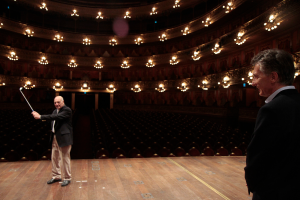 Ein älterer Mann in einem Anzug hält einen Golfschläger auf der Bühne, neben ihm steht ein weiterer älterer Mann rechts, unter einer Decke mit vielen Sitzplätzen darunter.