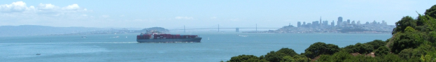 Ein großer Containerfrachter fährt durch Wasser in der Nähe einer Stadt, mit einer Brücke im Hintergrund, Bäumen auf der rechten Seite, Gebäuden in der Ferne und Wolken am Himmel.