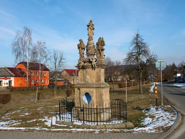Ein Holocaust-Gedenkdenkmal in einer Kleinstadt, umgeben von einem Metallzaun, mit Bäumen, Häusern, Fahrzeugen, Strommasten und einem bewölkten Himmel im Hintergrund.