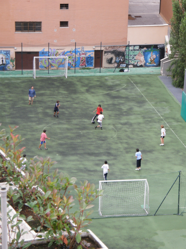 Kinder beim Fußballspielen mit Toren, ein Gebäude mit Graffiti im Hintergrund, Pflanzen links und Bäume rechts.