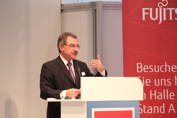 Eine Person mit Brille, Blazer und Krawatte steht neben einem Rednerpult mit Mikrofon und Maus, mit einer Wand und einem Banner im Hintergrund.