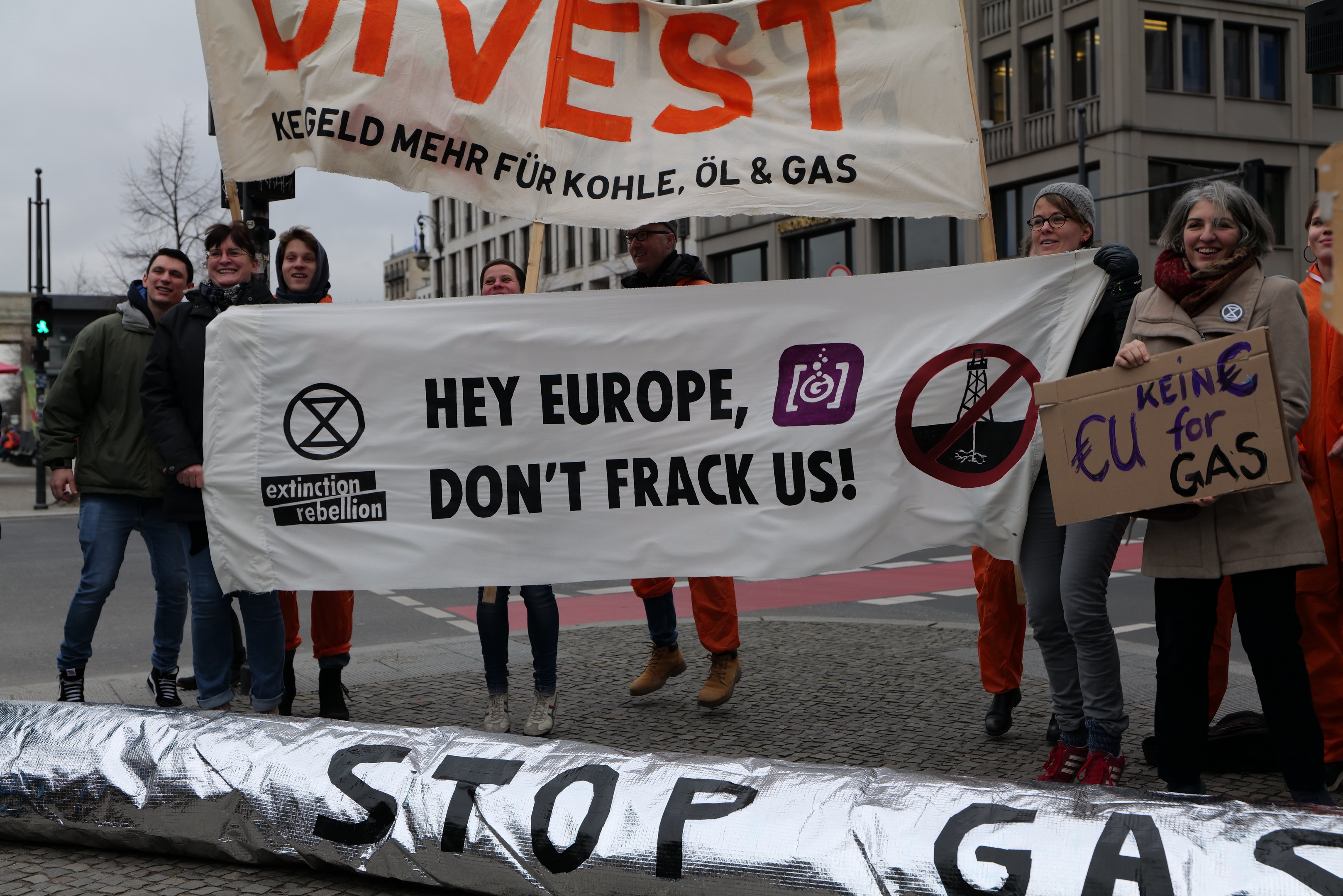 Gruppe von Menschen auf einer Straße mit einer 'Stoppt Gas'-Plakette protestieren gegen die Europäische Union, mit Gebäuden, Bäumen, Pfählen, Verkehrszeichen und einem klaren blauen Himmel im Hintergrund.