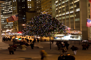 Ein Weihnachtsbaum auf einer Stra√Ÿe mit Menschen drumherum, flankiert von zwei Fahnenmasten, mit beleuchteten Geb√§uden im Hintergrund.