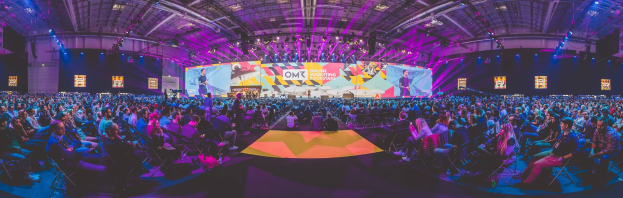 Eine große Menschenmenge sitzt vor einer hell erleuchteten Bühne mit mehreren stehenden Personen, wahrscheinlich bei einem Online-Marketing-Event.