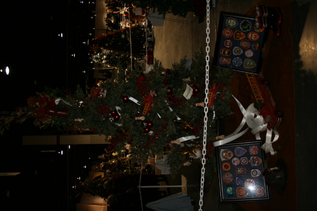 Ein geschmückter Weihnachtsbaum mit Geschenken, Rahmen und Spielzeugen auf einer Plattform, mit Menschen, zusätzlichen Weihnachtsbäumen und Lichtern im Hintergrund.