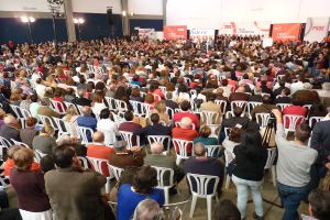 Volles Stadion mit vielen sitzenden und stehenden Menschen, Bühne mit zwei Rednern am Podium und zahlreichen Bannern und Lichtern.