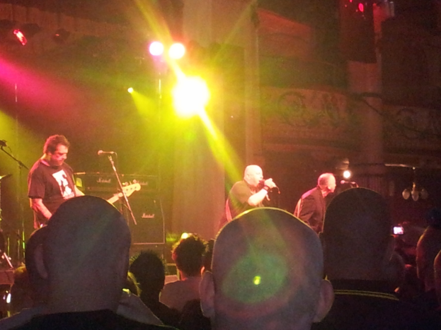 Drei Männer musizieren auf der Bühne bei einem Konzert, mit einem Gitarristen in einem schwarzen T-Shirt links, einer großen Menge unten und Lichtern und einem Gebäude im Hintergrund.