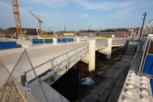 Eine Baustelle mit einer Brücke über Wasser, umgeben von Gebäuden, Kränen, Straßenlaternen, Strommasten, Stromkabeln, Kraftfahrzeugen, Containern, Bäumen und einem bewölkten Himmel.