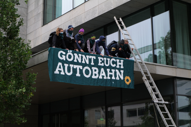 Eine Gruppe von Menschen auf einem Gebäude, das eine Fahne hält, mit einem Baum links und Glasfenstern im Hintergrund, wahrscheinlich protestierend in Unterstützung der deutschen Regierung.