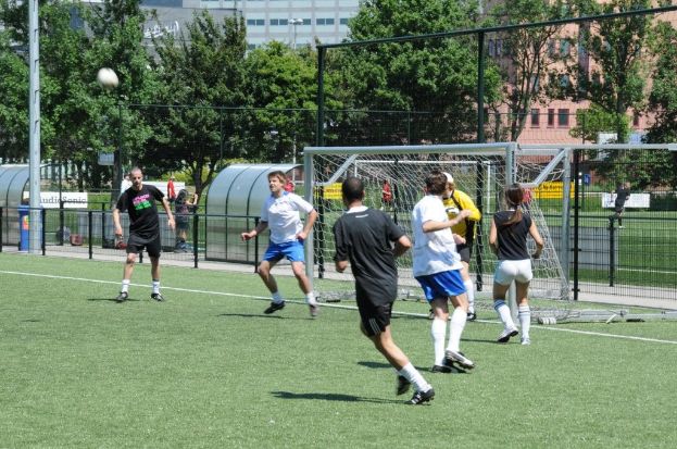 Menschen beim Fussballspielen auf einem Feld mit einem Zaun und einem Tor in der Mitte, einem Pfahl und Ball in der oberen linken Ecke und Bäumen und Gebäuden im Hintergrund.