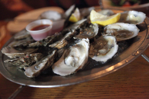 Ein Teller mit mehreren Muscheln nebeneinander, eine Tasse Sauce in der Mitte, zwei Zitronenstücke daneben und Teller im Hintergrund auf einem Tisch.