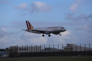 Germanwings Airbus A320-200 beim Start von Frankfurt Airport, mit Gras darunter, einem Zaun im Vordergrund, Gebäuden und Bäumen im Hintergrund und einer bewölkten Himmel überhaupt.