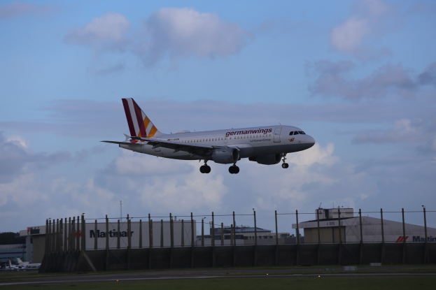 Germanwings Airbus A320-200 beim Start von Frankfurt Airport, mit Gras darunter, einem Zaun im Vordergrund, Gebäuden und Bäumen im Hintergrund und einer bewölkten Himmel überhaupt.