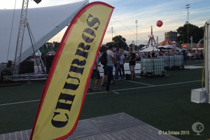 Eine lebhafte Karnevalsszene mit Zelten, einem zentralen Mast, Menschen, Bäumen, Strommasten, Straßenbahnen auf der Straße und einem Eisenständer auf der linken Seite unter einem klaren Himmel.