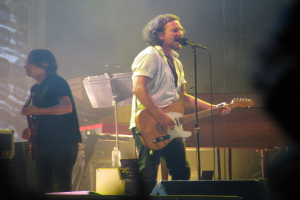 Ein Mann in einem weißen Hemd spielt Gitarre und singt in ein Mikrofon auf der Bühne, neben ihm ein Mann in einem schwarzen Hemd, der ebenfalls Gitarre spielt; im Hintergrund sind ein Piano, Kabel, Lautsprecher und Lichter zu sehen.
