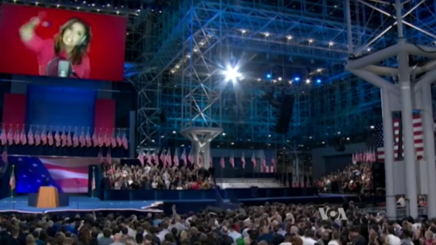 Eine große Menschenmenge sitzt einer Bühne mit einem Podium und einem Mikrofon gegenüber, Fahnen im Hintergrund, ein Bild einer Frau auf einem Bildschirm und Lampen und Stangen oben, was auf eine Präsidentschaftsdebatte hindeutet.