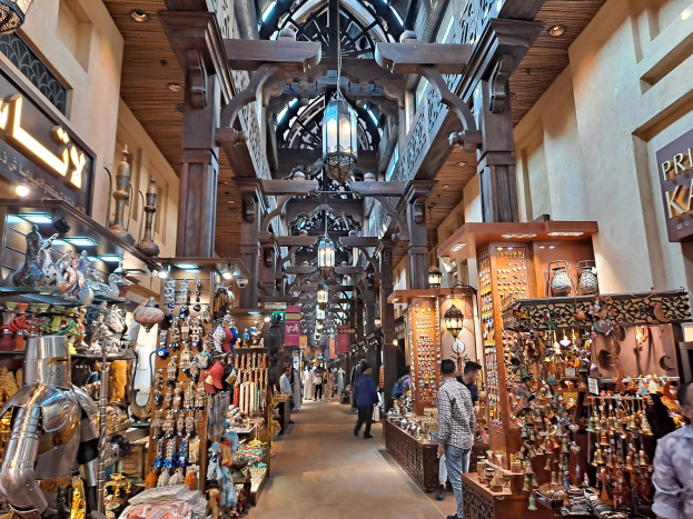 Ein belebter Dubai-Basar mit Menschen, die zwischen Regalen voller Waren hindurchgehen, Wänden mit Texttafeln und einer Decke mit warmem Licht.