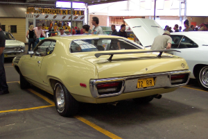 Ein Oldtimer ist auf einer Stra├če mit Menschen in der N├Ąhe geparkt, ein Laden in der Mitte und Geb├Ąude im Hintergrund.