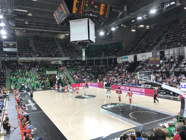 Basketballspiel in einer großen Arena mit Zuschauern, mit Beteiligung der Teams Asvel und Galatasaray, unter Stadionbeleuchtung mit Anzeigetafel.