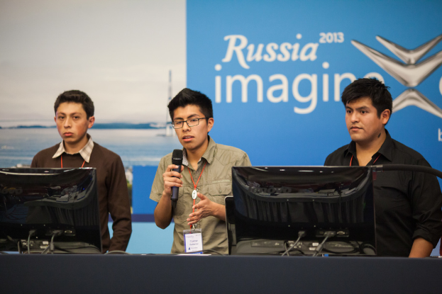 Drei Jungen stehen neben einem Schreibtisch mit zwei Computersystemen, einer spricht in ein Mikrofon, tragen ID-Karten mit roten Tags, vor einer Wand mit einer Werbetafel, auf der 'Russia Imagine 2013' steht.