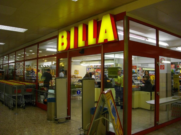 Ein Supermarkt mit einem Eingang in der Mitte, Mülltonnen daneben, einer Reihe Einkaufswagen auf der linken Seite und einigen Menschen drinnen, die unter Deckenbeleuchtung Produkte durchstöbern.