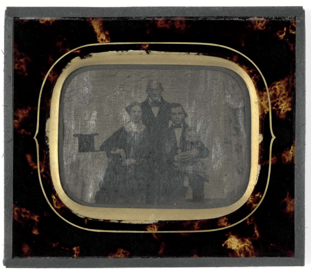 Schwarze-und-weiß-Foto eines Mannes in Anzug und Krawatte und einer Frau in einem unbekannten Outfit, die vor einem alten Fernseher stehen, beide mit ernsten Gesichtern.