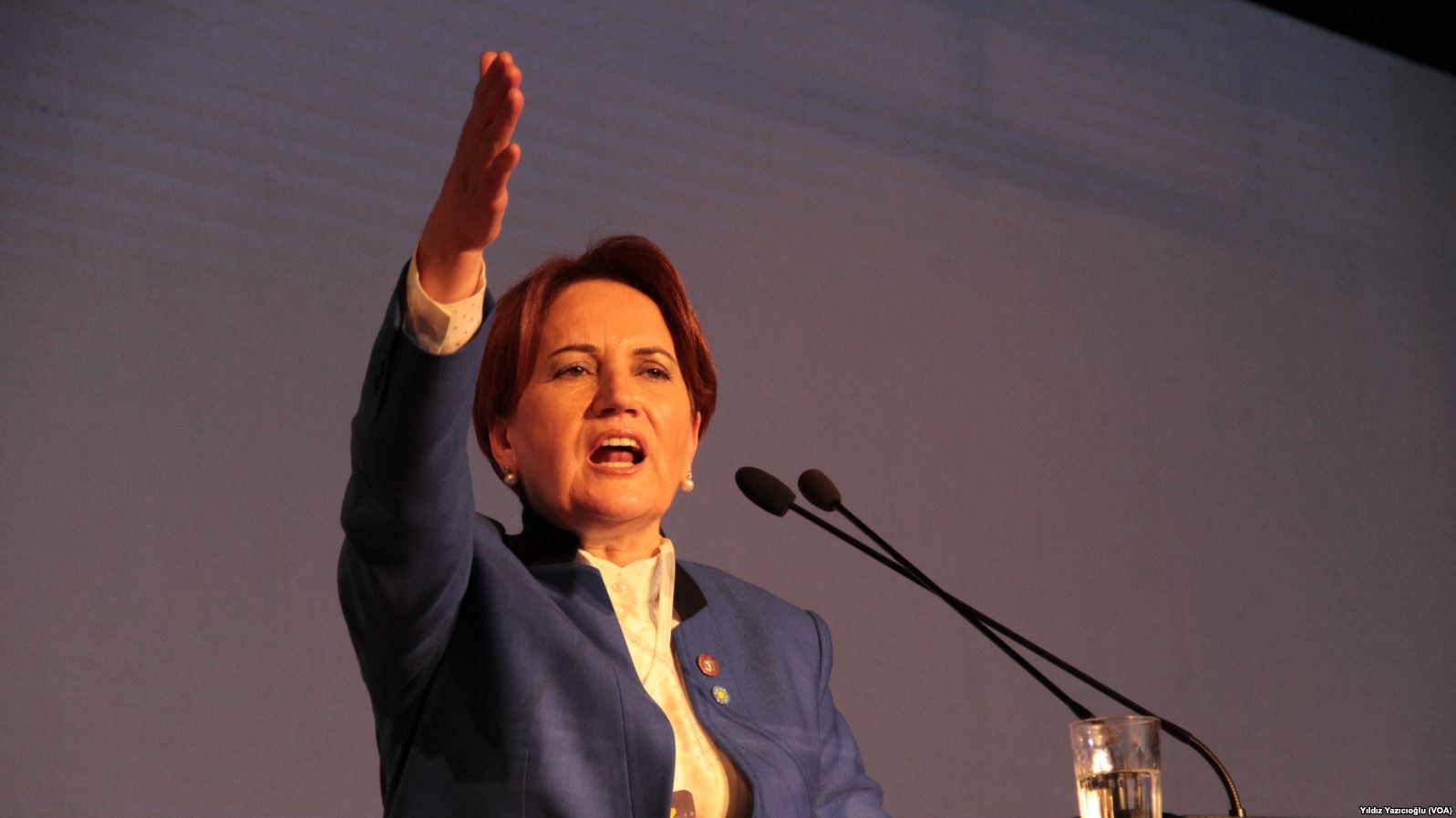 Frau steht an einem Rednerpult mit einem Bildschirm dahinter und hält eine Rede, während sie ein Glas Wasser und zwei Mikrofone auf dem Pult hat.