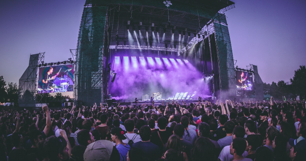 Eine große Menge bei einem Musikfestival schaut auf einer hell erleuchteten Bühne mit Instrumenten und Bildschirmen, die vor einem Gebäude, Bäumen und einem klaren blauen Himmel steht.