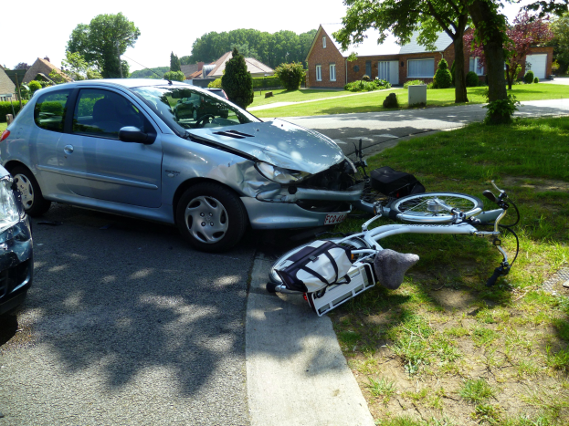 Ein Auto ist auf der Straße gegen ein Fahrrad geprallt, das auf dem Gras daneben liegt, Häuser und Bäume im Hintergrund unter einem klaren blauen Himmel.