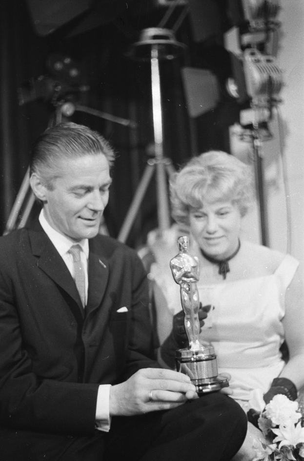 Ein Mann in einem schwarzen Blazer, weißem Hemd und Krawatte hält eine Oscar-Statue in der Hand und sitzt neben einer Frau, die einen Blumenstrauß hält, beide in Abendgarderobe, mit Scheinwerfern im Hintergrund, in Schwarz-Weiß.