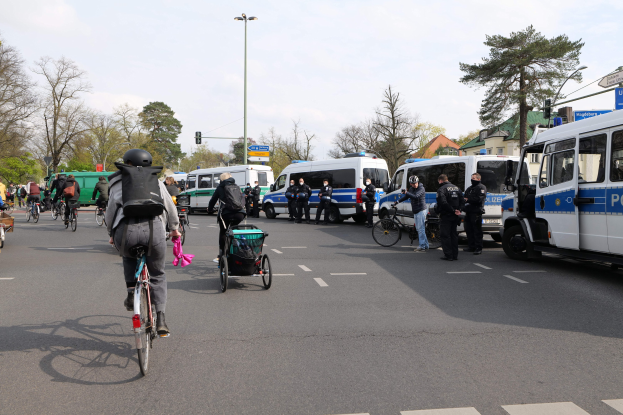 Eine Gruppe von Menschen, die auf Fahrrädern eine Straße entlangfährt, mit Polizeiwagen und anderen Fahrzeugen, einige tragen Helme und haben Taschen oder schieben Kinderwagen, mit Bäumen, Gebäuden und Verkehrszeichen im Hintergrund.