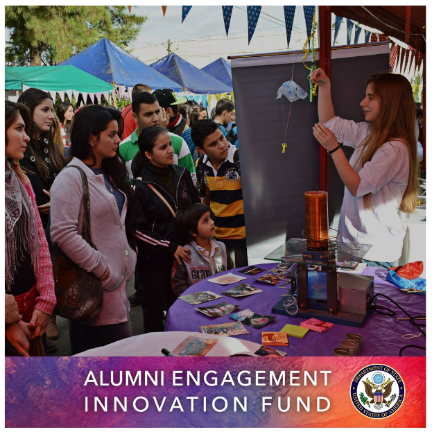 Gruppe von Menschen um einen Tisch versammelt mit einem "Alumni Engagement Innovation Fund"-Schild, umgeben von Gegenständen, mit Zelten und Bäumen im Hintergrund unter einem klaren Himmel, einschließlich Text und Logo unten.