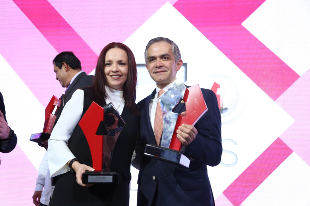 Zwei Personen auf einer Bühne, die lächeln und Trophäen halten, mit einem sichtbaren Bildschirm im Hintergrund.