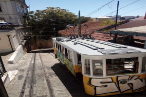 Eine gelbe und weiße Tram auf einer Schiene in Lissabon, Portugal, mit Gebäuden auf beiden Seiten und Bäumen und einem klaren blauen Himmel im Hintergrund.