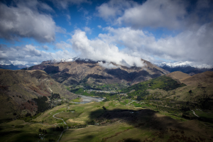 Ein atemberaubender Ausblick von der Spitze eines Berges in Queenstown, Neuseeland, der grüne Wiesen, Bäume, eine kurvenreiche Straße und einen Himmel mit weißen, flauschigen Wolken zeigt.