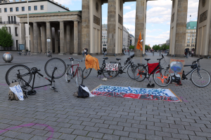 Eine Gruppe von Fahrr├Ądern vor dem Brandenburgertor in Berlin geparkt, mit Taschen und einem Banner in der N├Ąhe, vor einer Kulisse aus Geb├Ąuden, B├Ąumen, Masten, Flaggen und einem klaren blauen Himmel.