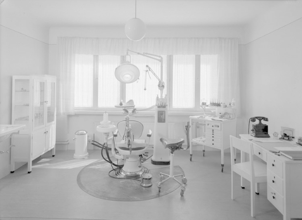 Ein Schwarz-Weiß-Foto einer Zahnarztpraxis mit einem Stuhl, einem Tisch, einem Schrank, einem Waschbecken, einem Müllbehälter, einem Fenster mit Vorhängen und Deckenleuchten sowie verschiedenen Gegenständen, die eine gemütliche und einladende Atmosphäre schaffen.