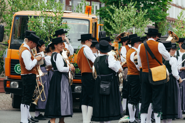 Eine Gruppe von Menschen in traditioneller bayerischer Tracht marschiert durch eine Straße und spielt Musikinstrumente, während im Hintergrund Bäume, Gebäude und ein geparktes Fahrzeug zu sehen sind.