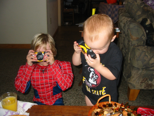 Zwei Kinder, eines im Spider-Man-Kostüm, halten eine Spielzeugkamera in der Nähe eines Holztisches mit einem Glas und einem Korb, mit einem Sofa auf der rechten Seite und einem weiteren Holztisch im Hintergrund.