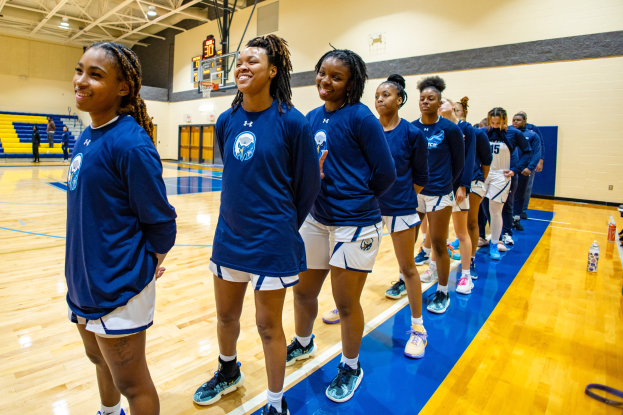 Eine Gruppe junger Frauen in blauen T-Shirts steht lachend auf einem Basketballfeld, mit Flaschen und Gegenständen rechts daneben und Zuschauern im Hintergrund.